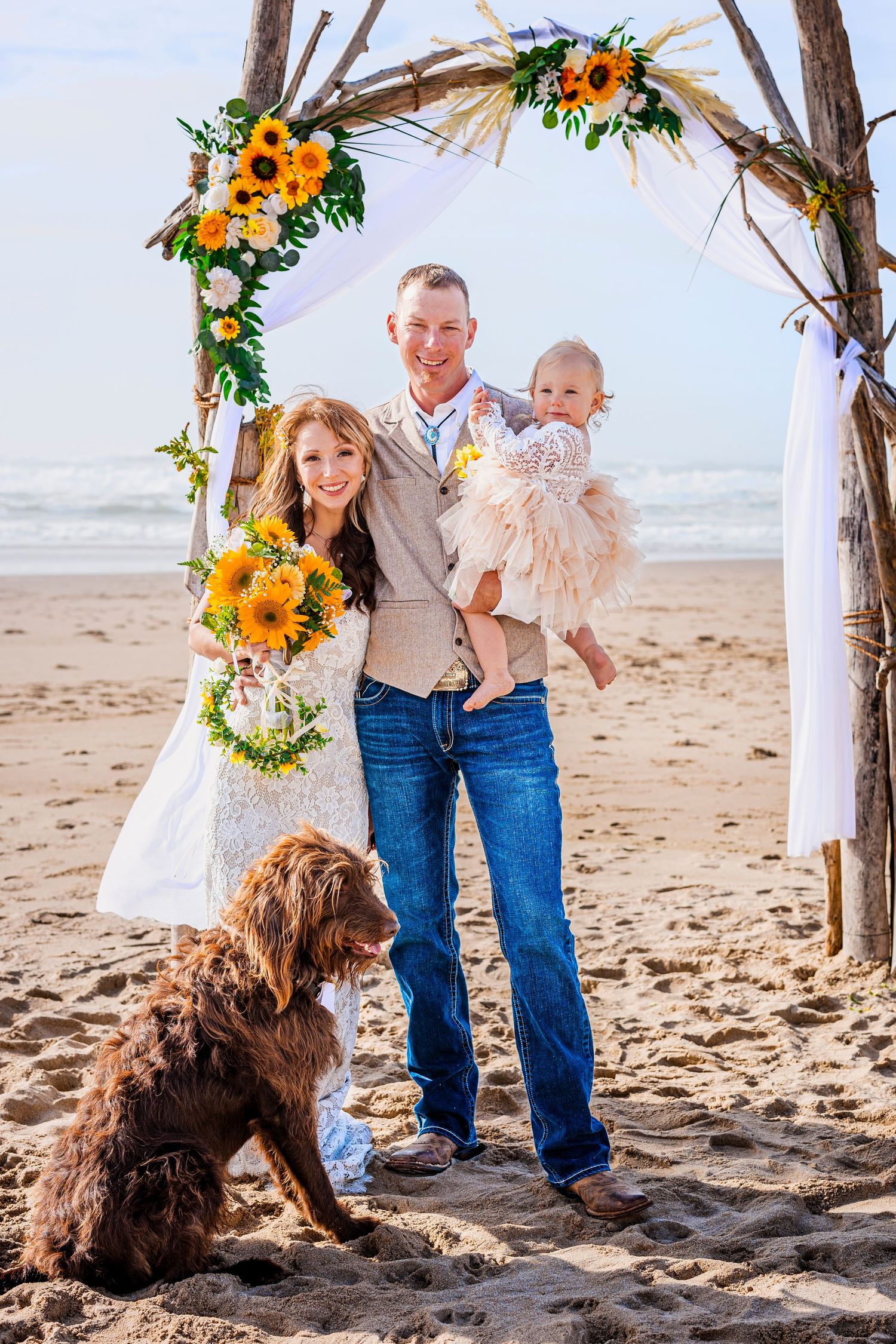 Seaside beach wedding ceremony setup with rustic arch decorated in sunflowers and white fabric.