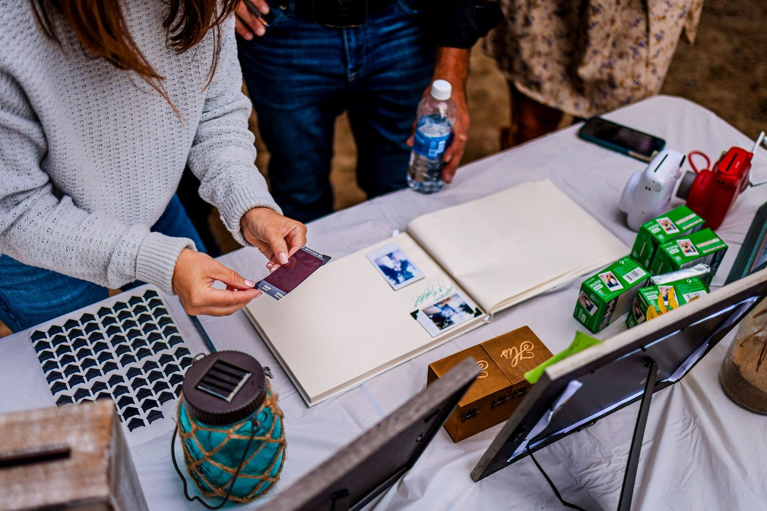 Hands work on papers at an outdoor table with a laptop, beverages, and toy cars visible.
