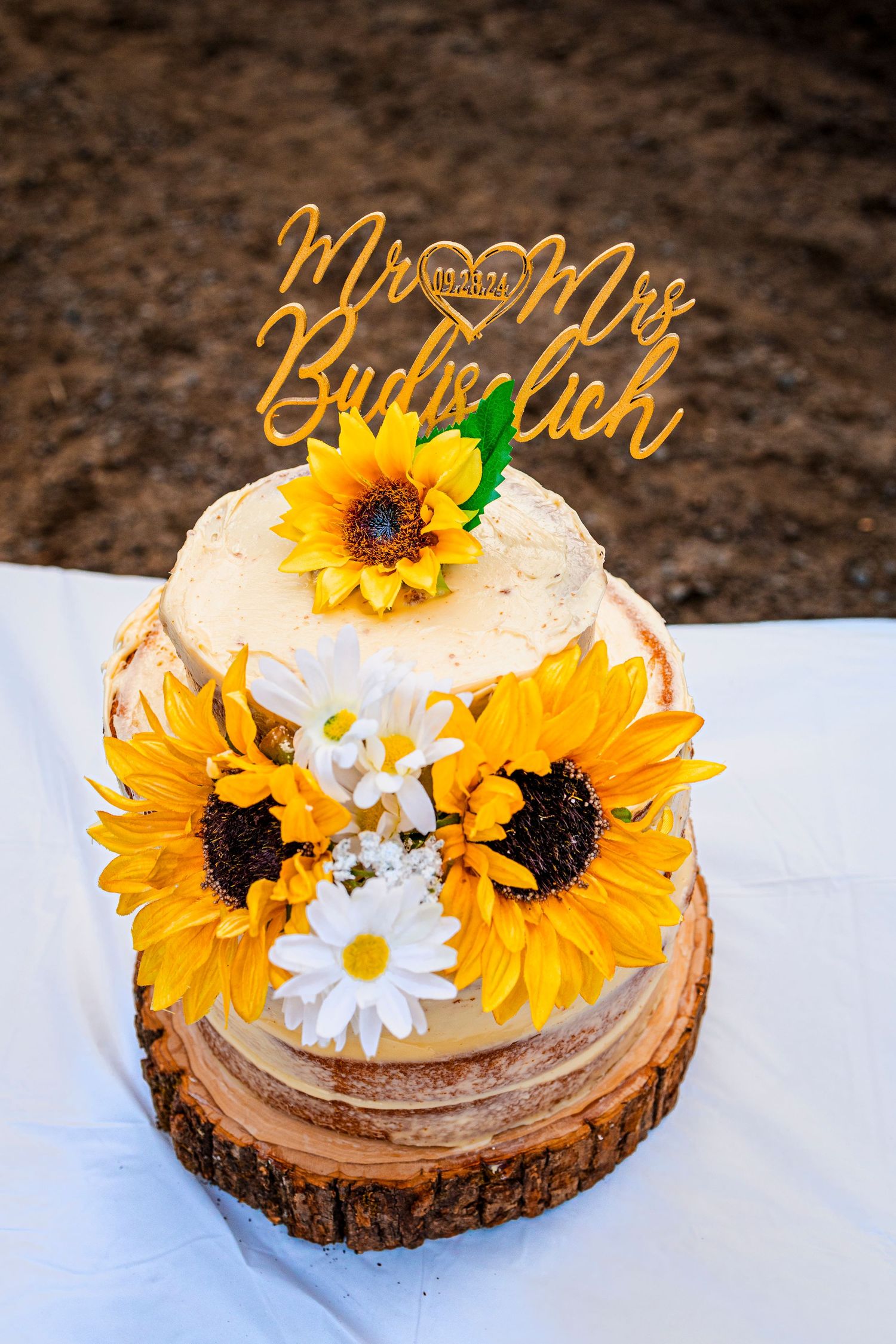 Rustic wedding cake decorated with bright sunflowers and daisies on a wood slice with a gold "Mr & Mrs" topper.