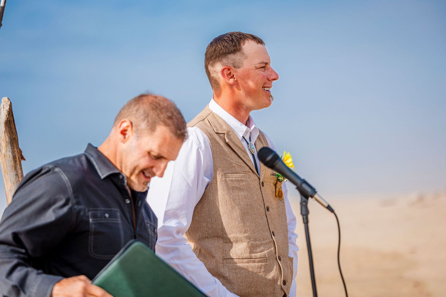 Two men sharing a laugh during an outdoor ceremony in a desert setting with microphone stand.