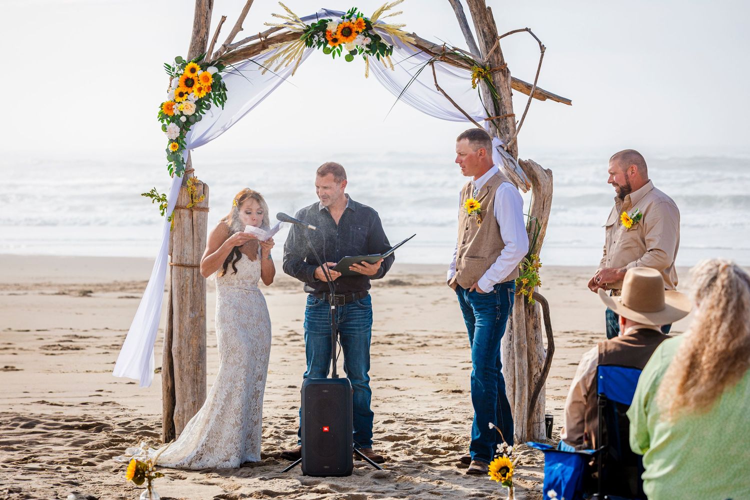A beachside wedding ceremony takes place under a rustic wooden arch decorated with orange flowers and white fabric.