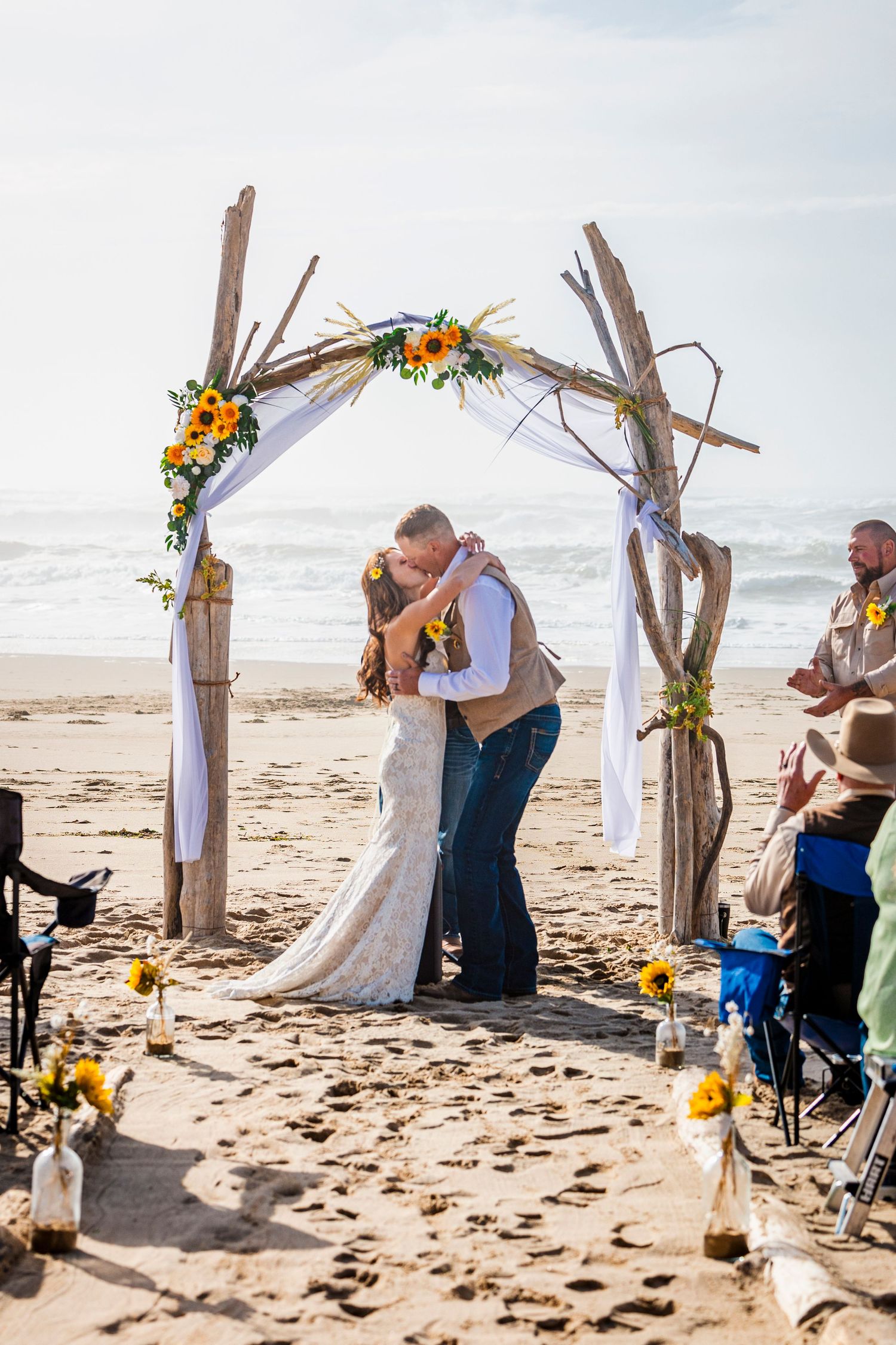 Intimate beach wedding ceremony under a driftwood arch adorned with sunflowers and flowing white fabric.