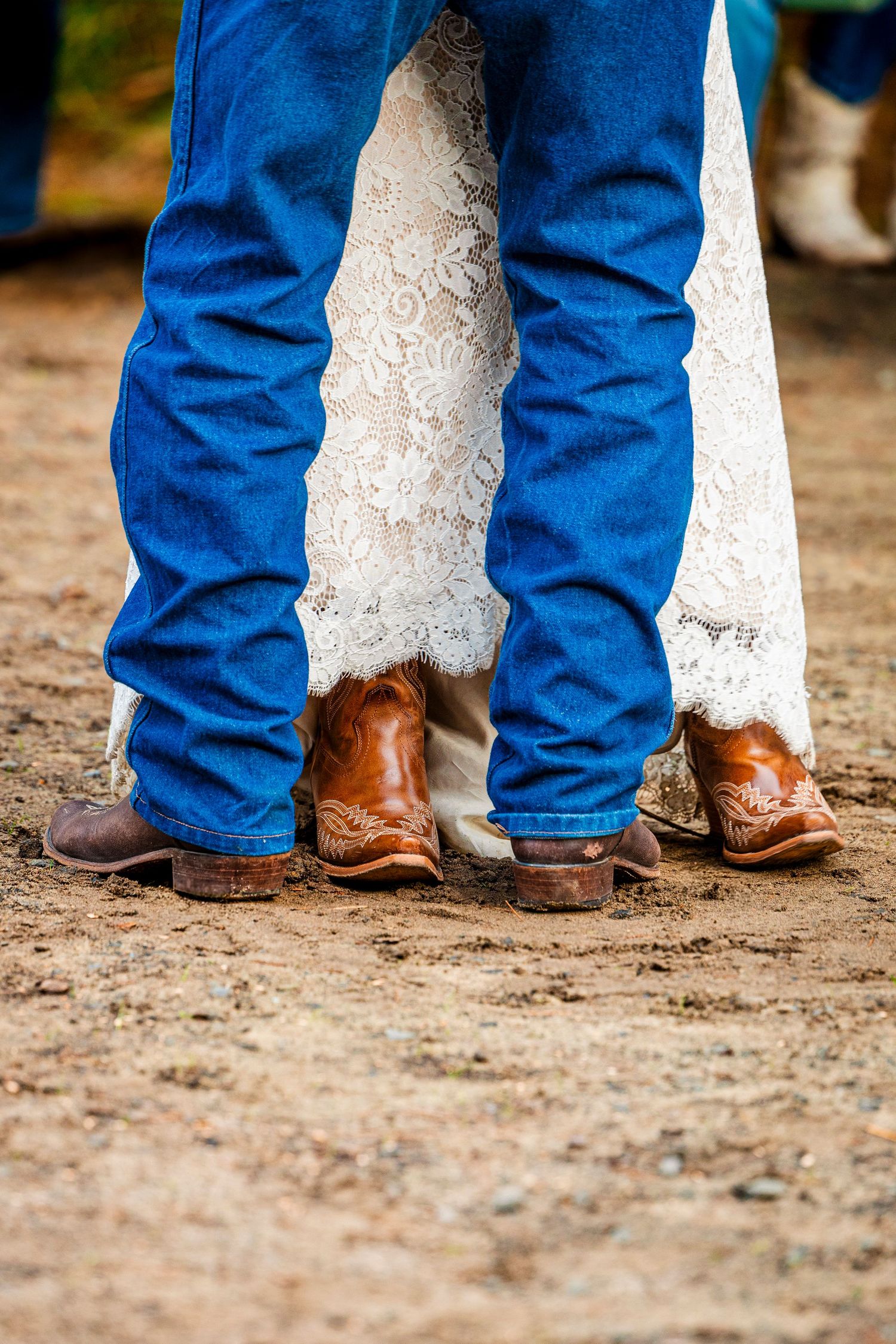 Close-up of cowboy boots and wedding dress hem on sandy ground.