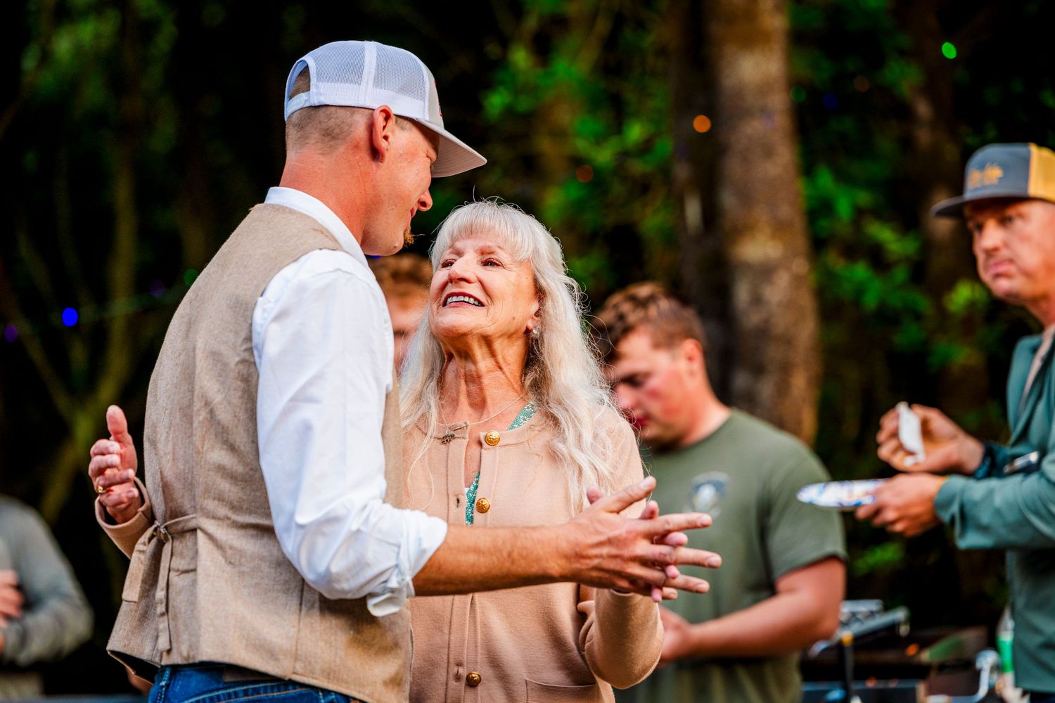 A joyful celebration moment between two people at an outdoor gathering with onlookers in the background.