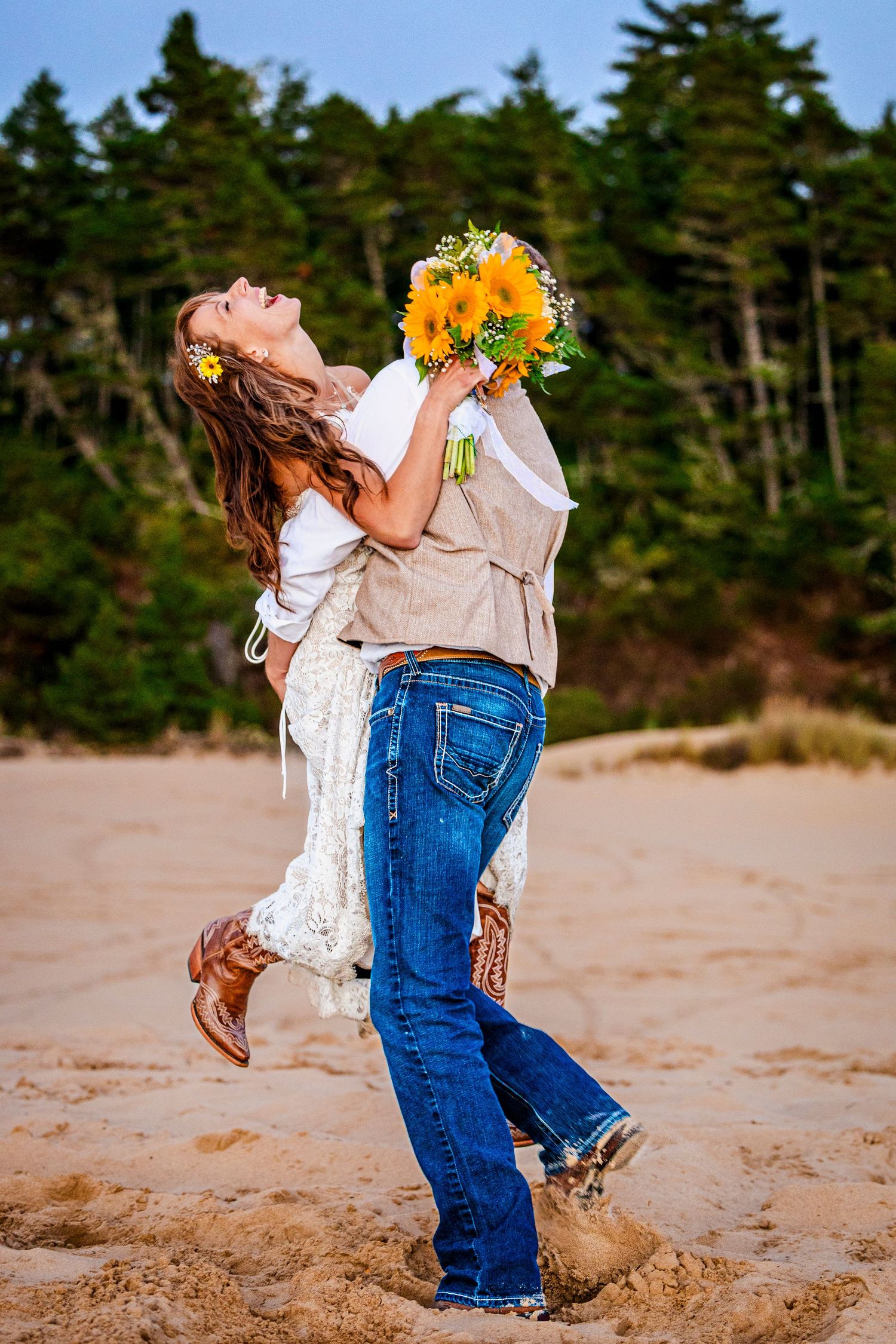 A joyful couple embraces on a beach with sunflowers and evergreen trees in background.