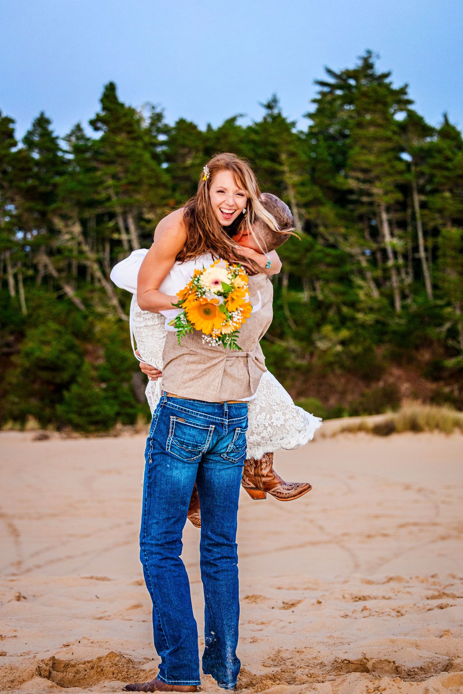 A couple embraces joyfully at the beach with sunflowers and pine trees in the background, the bride wearing a white dress.