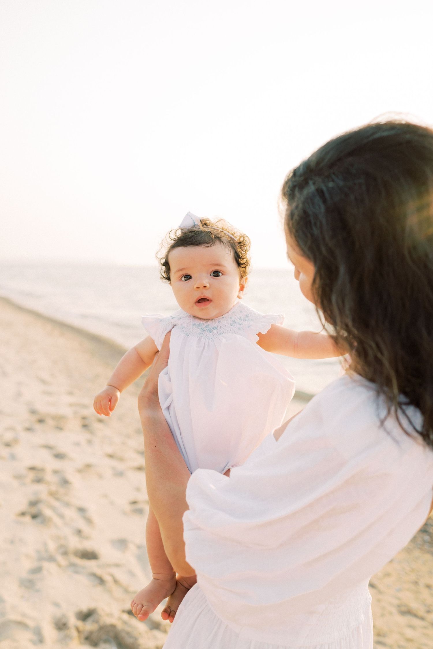 A baby wearing a white outfit is held on a peaceful beach at sunset while accompanied by an adult in white clothing.