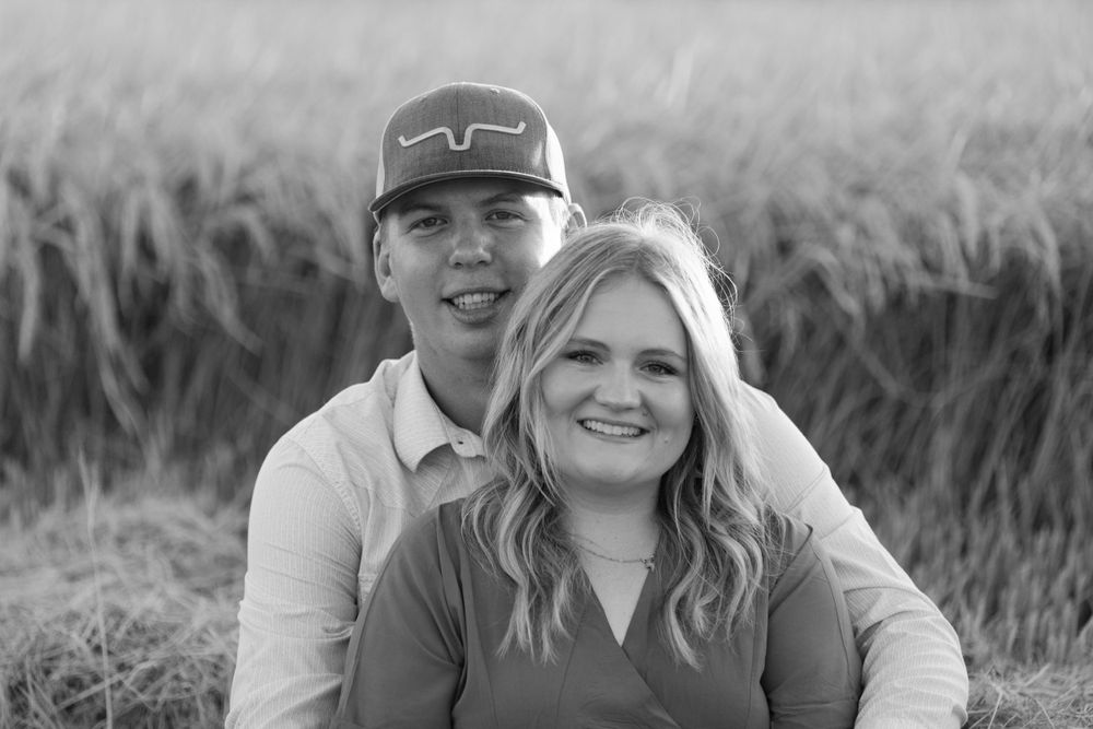 Black and white portrait of a smiling couple sitting together in front of a cornfield on a sunny day.