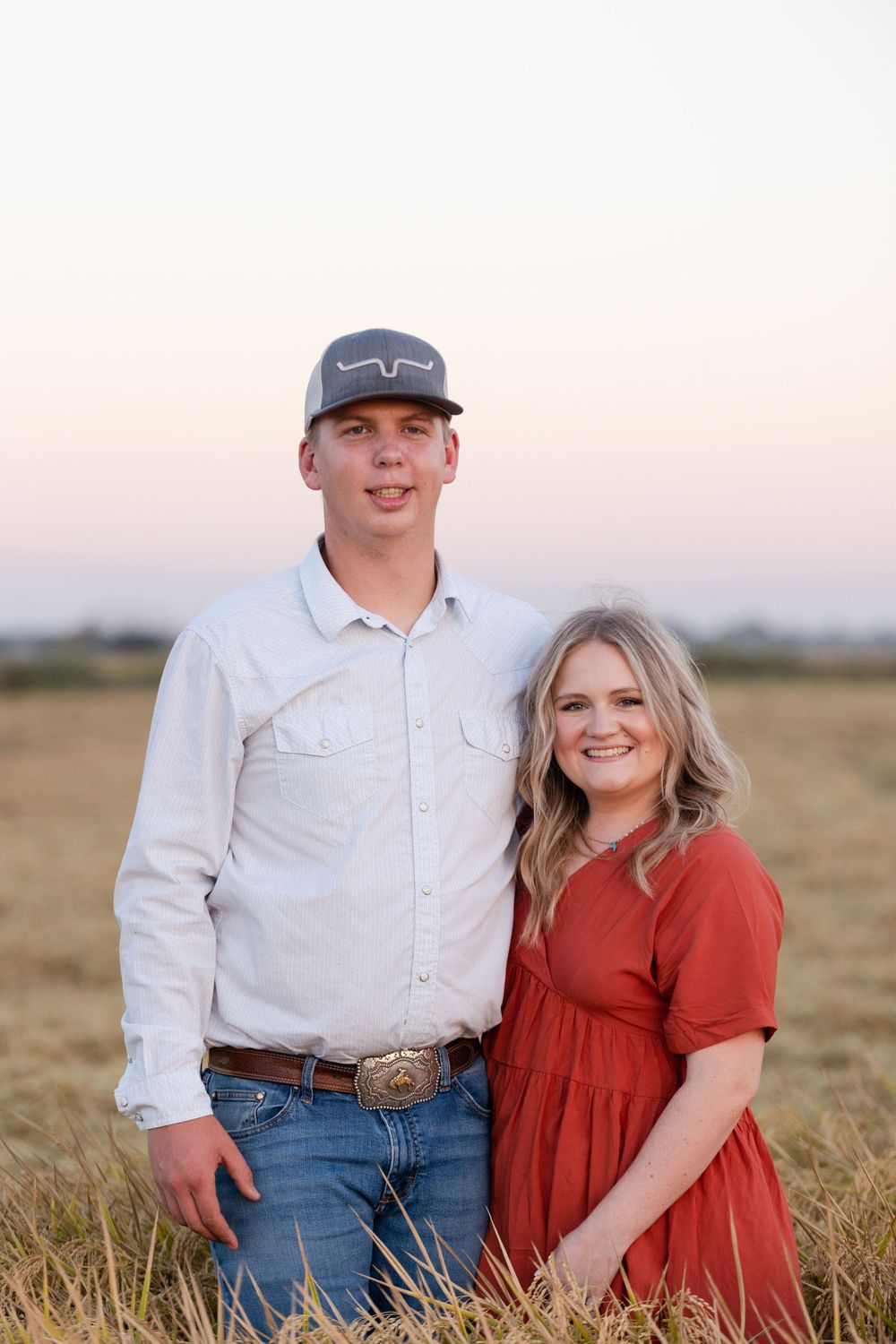 A couple poses together in a grassy field at sunset, wearing a white button-up shirt and rust-colored dress.