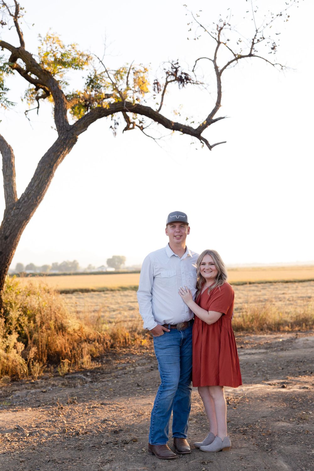 A couple poses together at sunset under a large tree in a rural field with golden light.