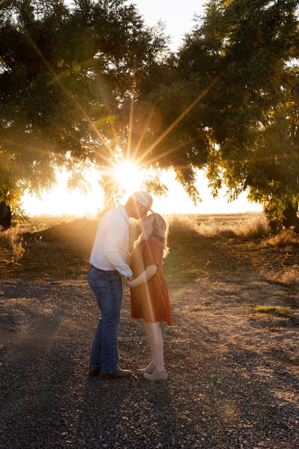 A couple embraces on a dirt path during golden hour sunset with sunburst rays shining through tree branches.