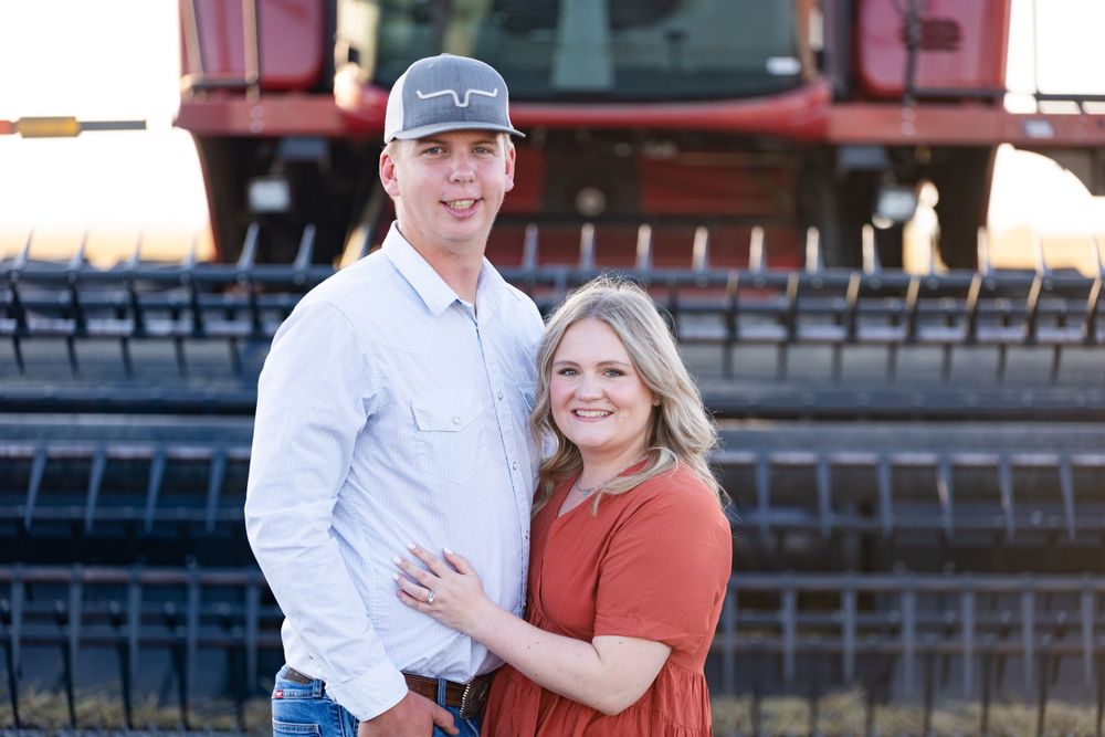 A couple poses together in front of a red combine harvester in a rustic farm setting.