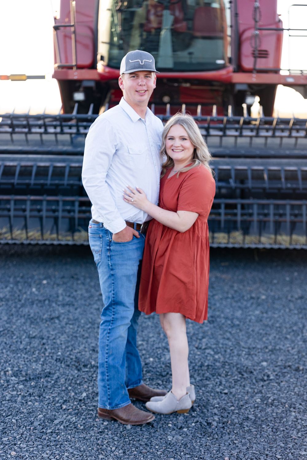 A couple poses in front of a red combine harvester in casual attire on a gravel surface.