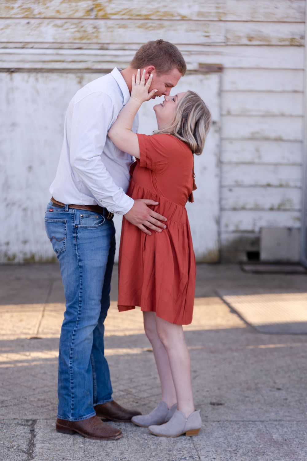 A romantic couple embraces in front of a rustic white wall, with the woman wearing a rust-colored dress.