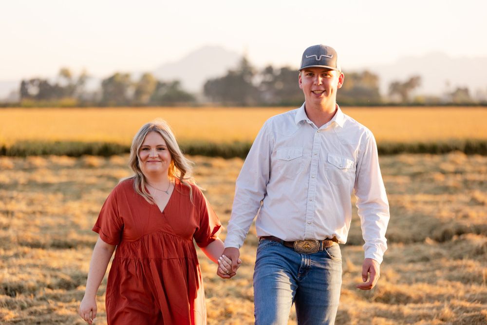 A couple walks hand in hand through a golden field during sunset with warm evening light behind them.