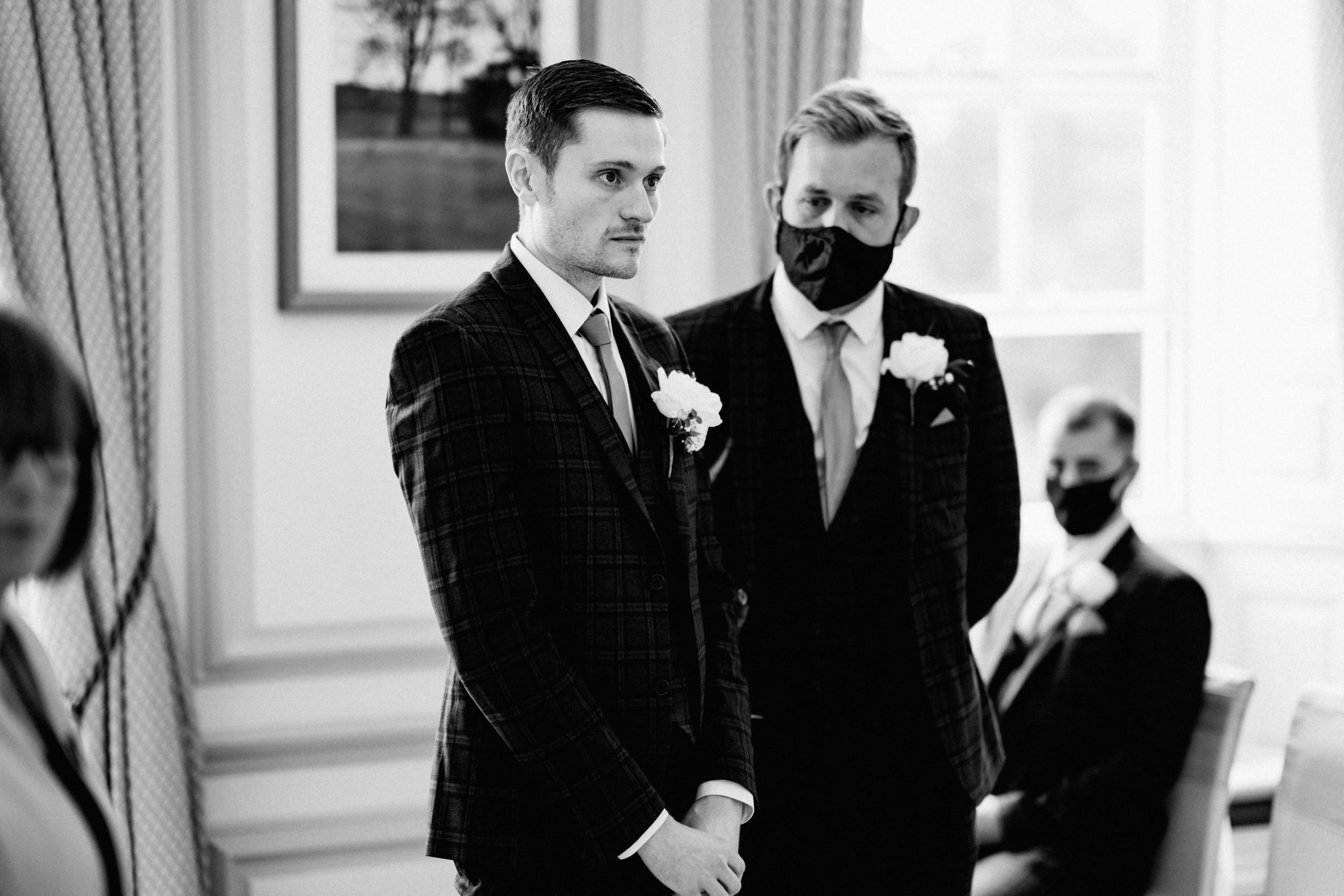 Two people in formal black suits wearing masks stand together during a wedding ceremony.