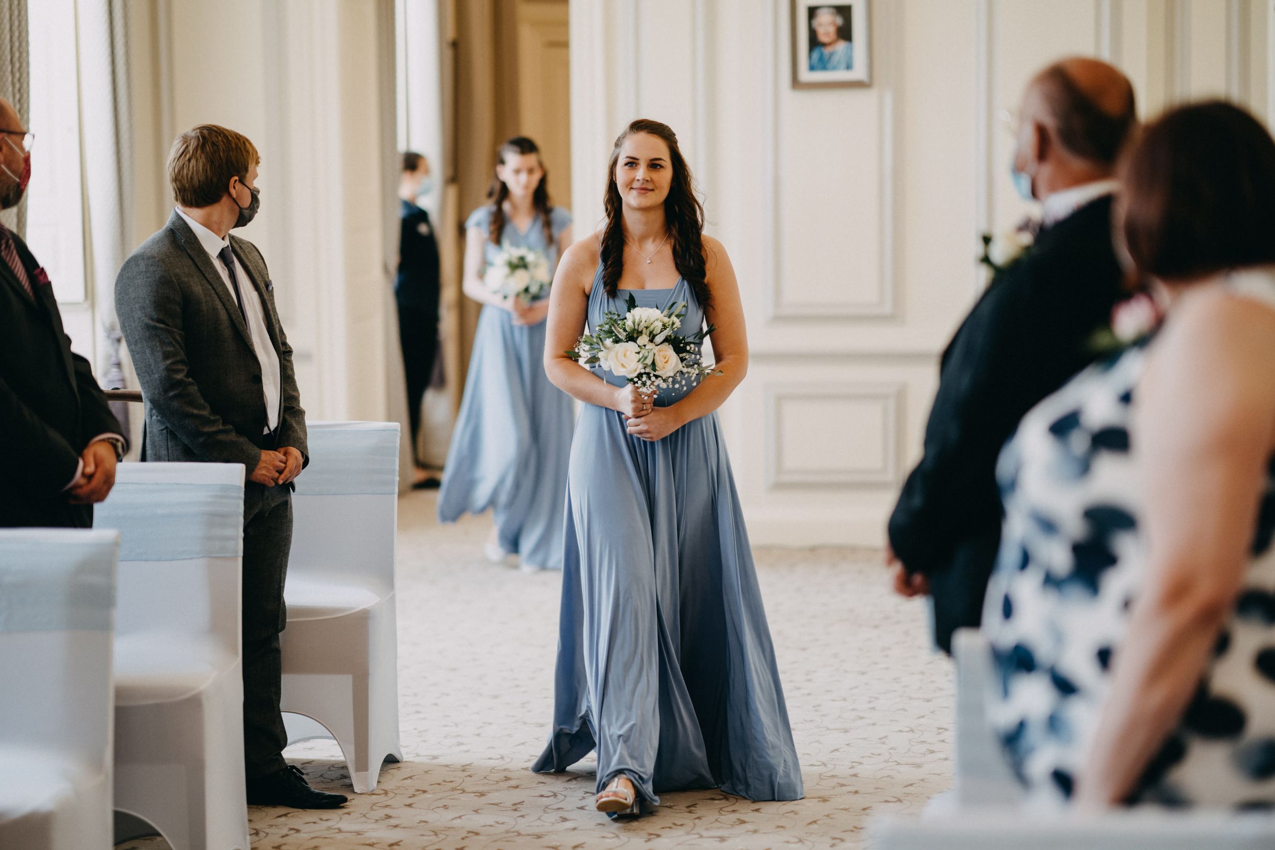 Bridesmaid in flowing light blue dress walks down aisle carrying bouquet.