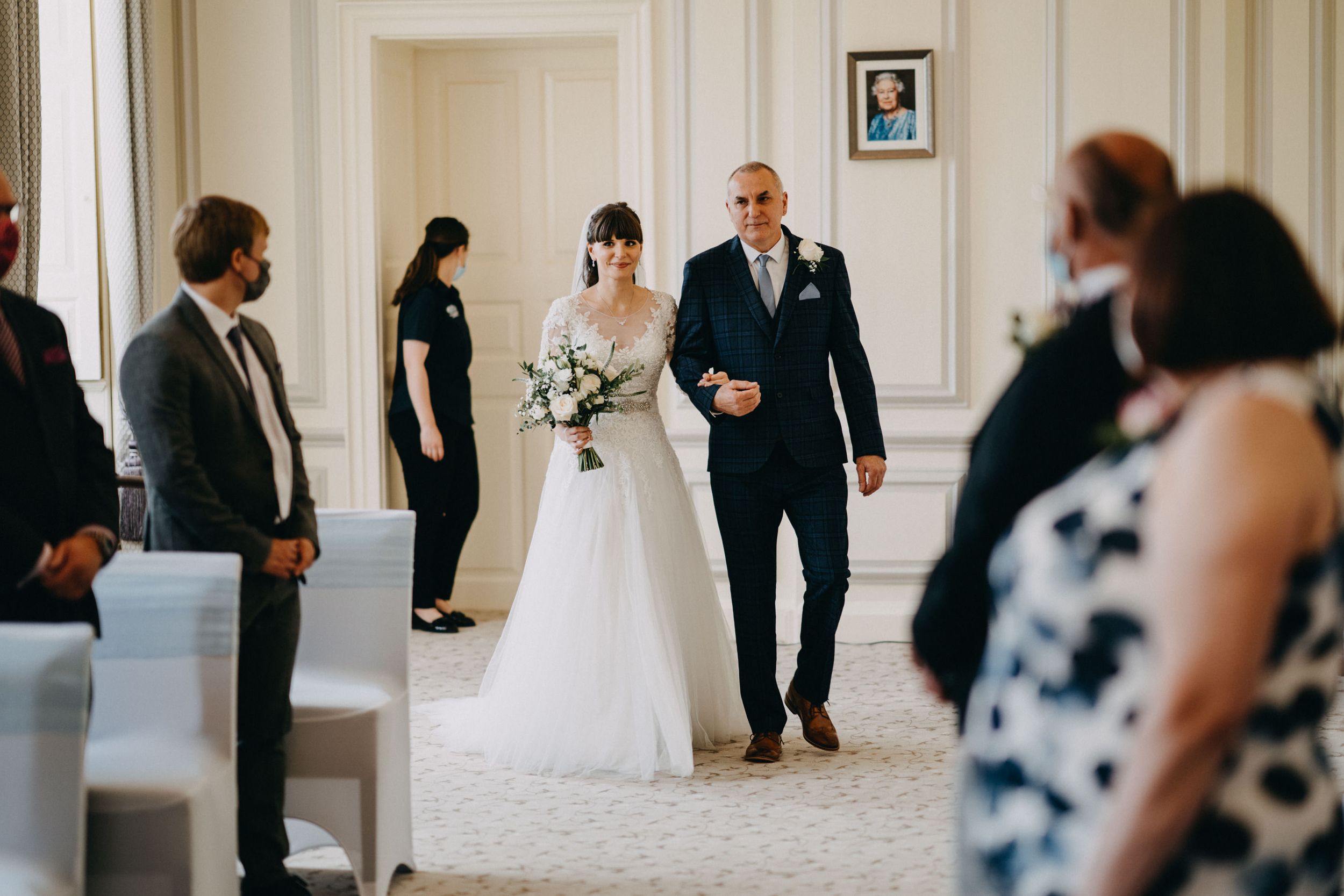 Wedding ceremony in elegant white room of Harrowden Hall with attendees watching processional.