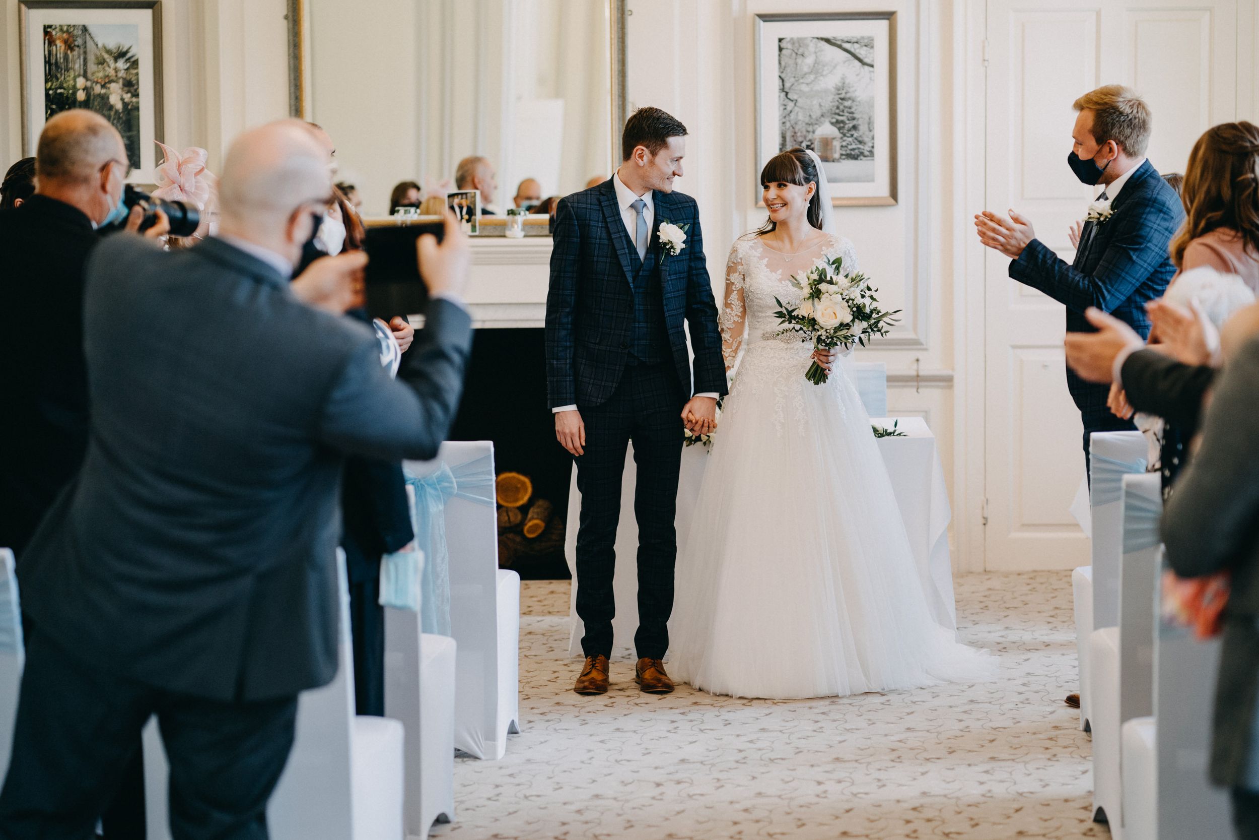 Wedding guests photograph a couple walking down the aisle at an elegant indoor ceremony venue.