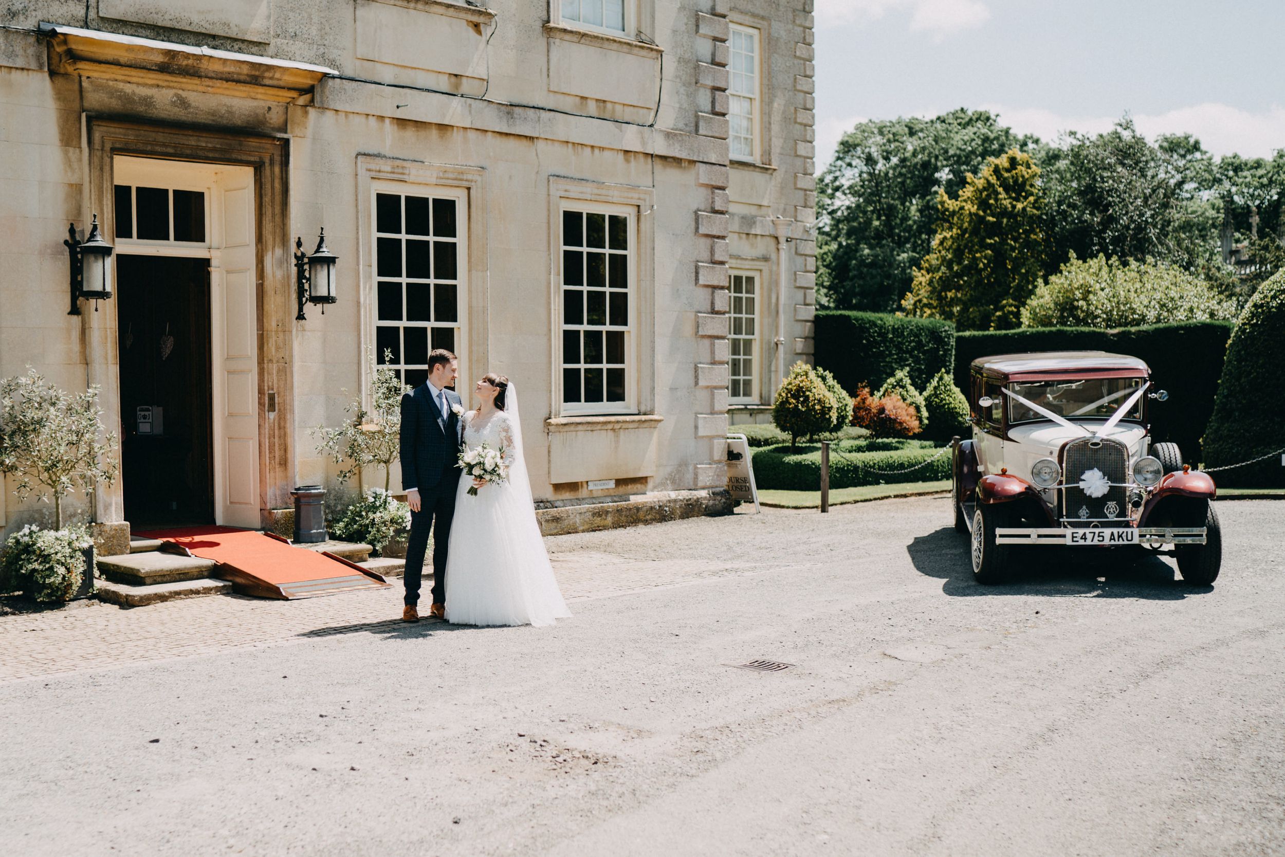 Newlyweds pose outside historic stone building of Harrowden Hall with classic vintage car.