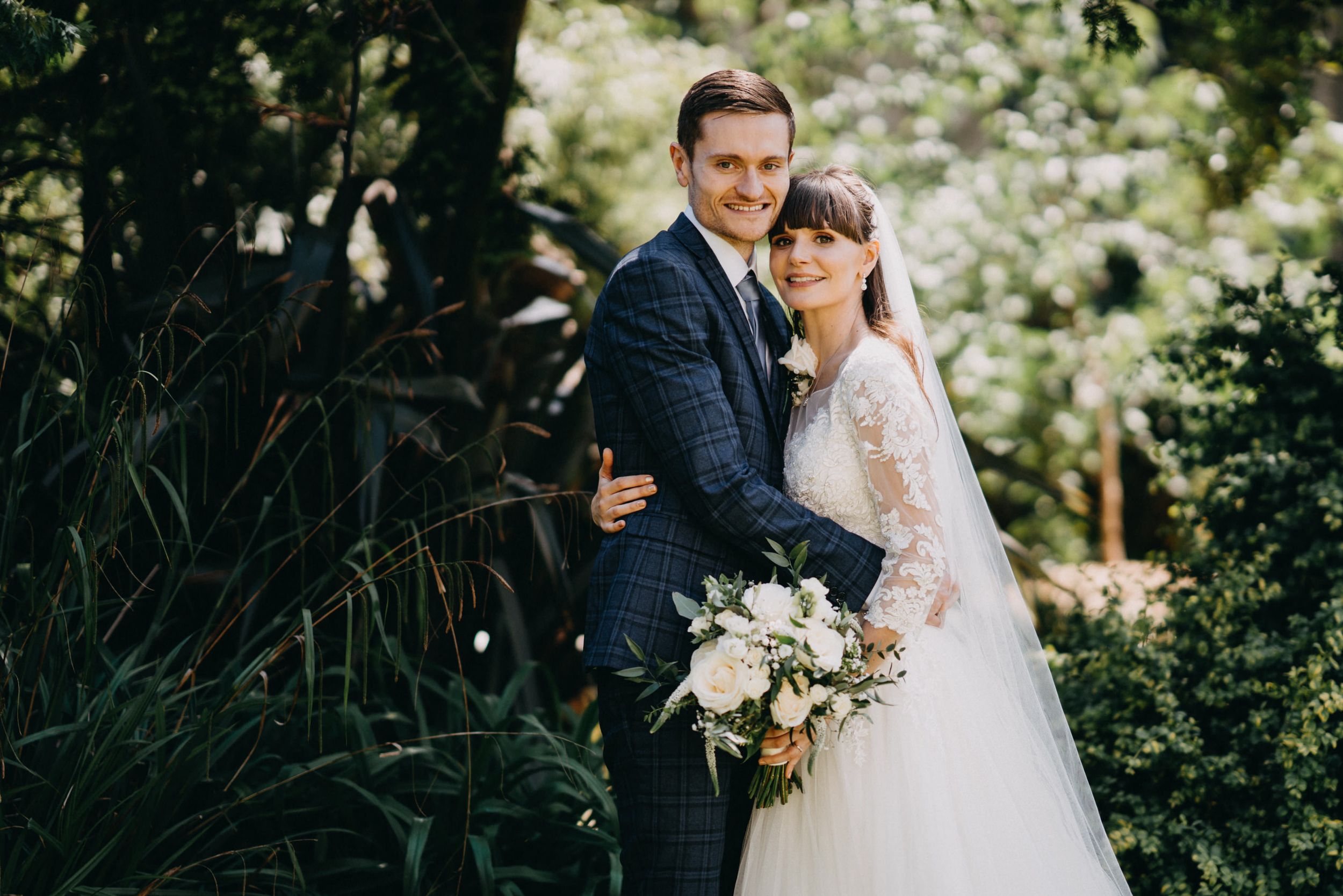 A romantic wedding portrait of a couple embracing outdoors with a white floral bouquet and lace dress details.