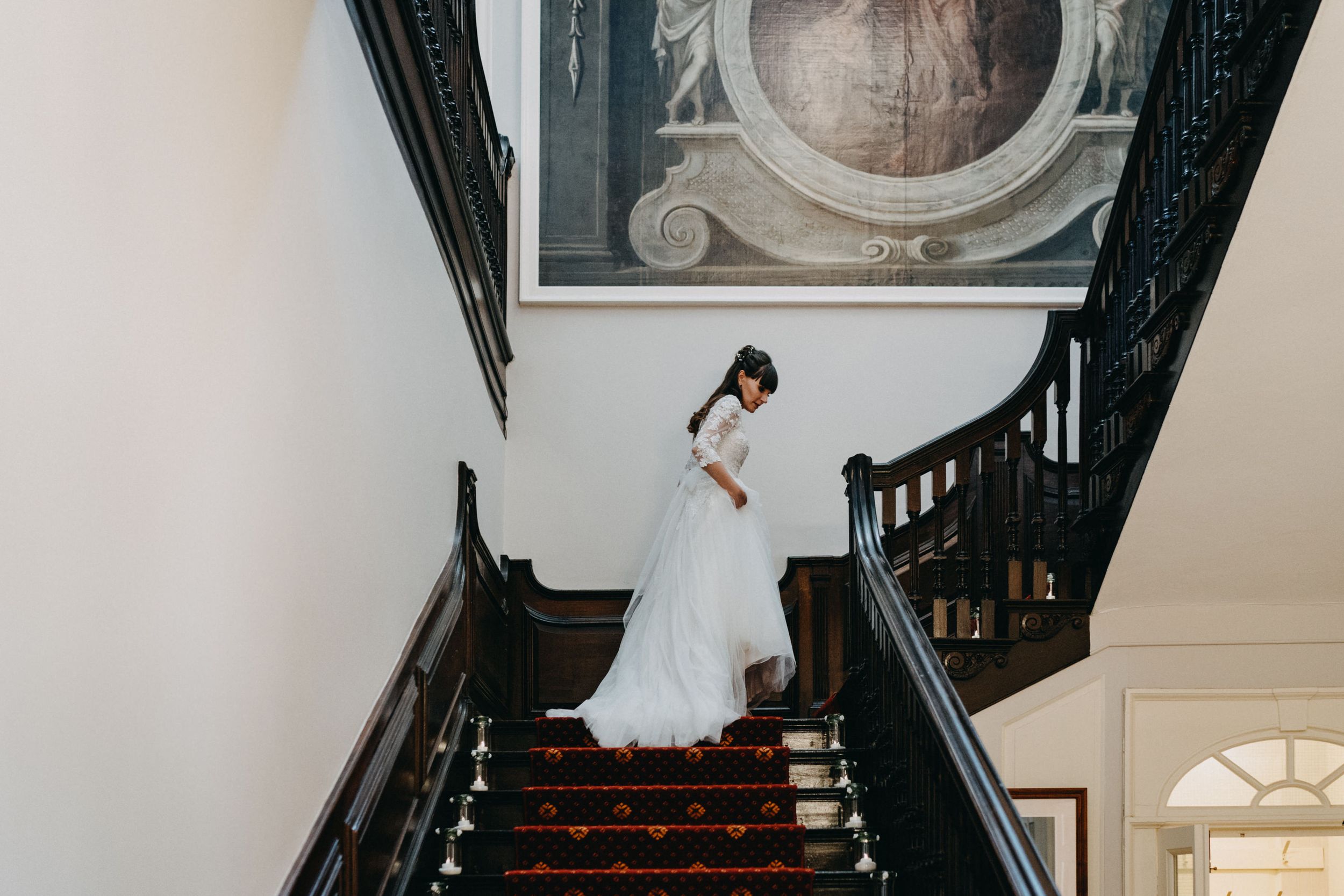 An elegant bride in a white gown descending a grand staircase with dark wood railings.