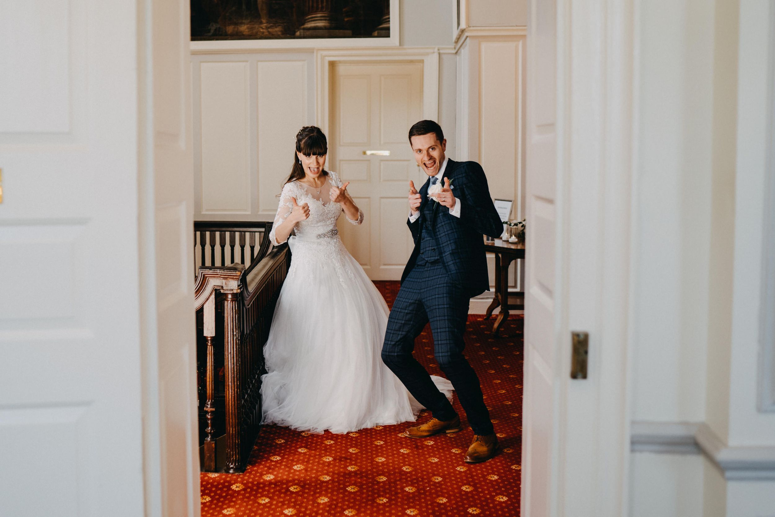 A playful wedding couple dancing in a hallway with vintage decor and red patterned carpet.