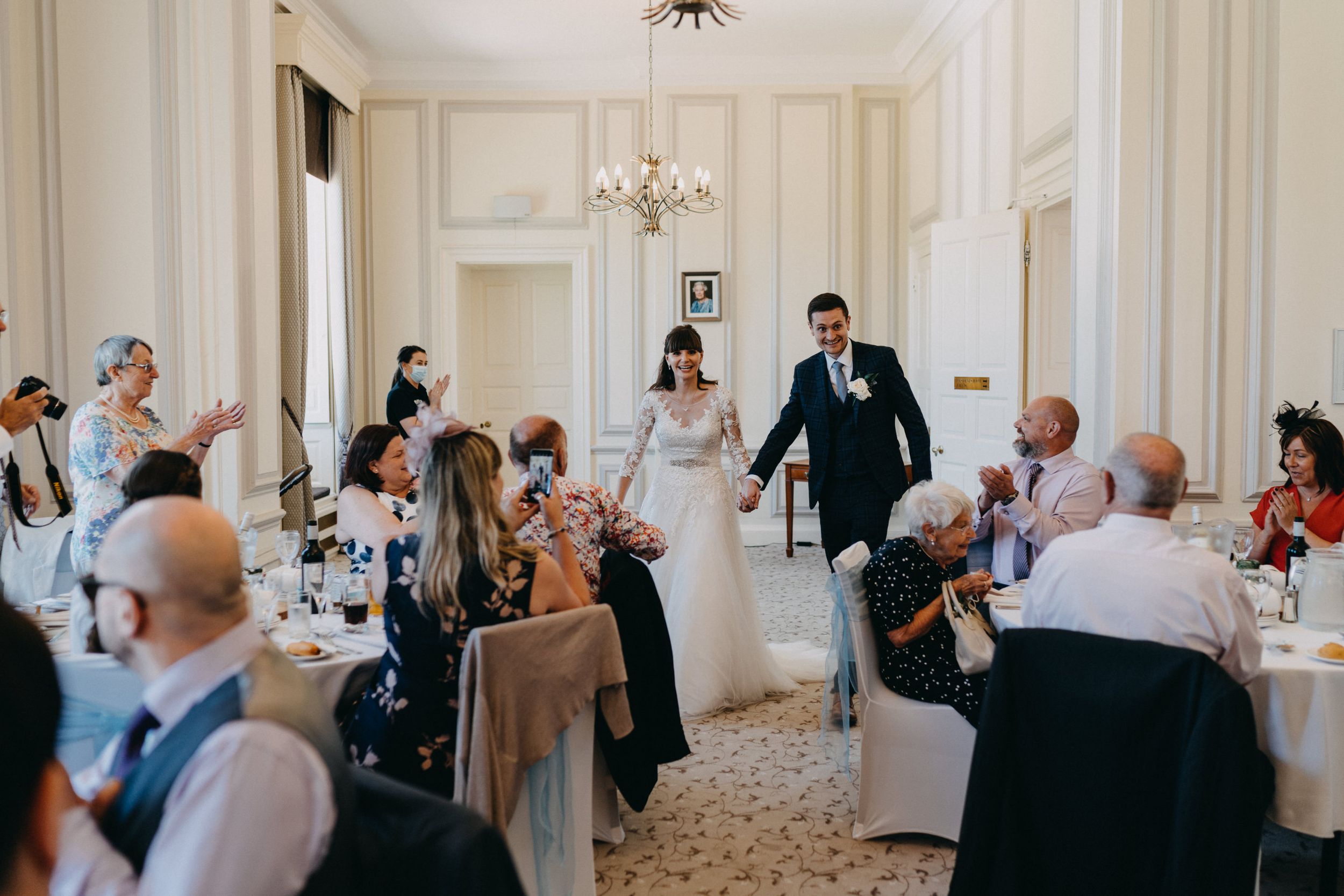 A wedding reception scene with guests seated at decorated tables in an elegant white room.
