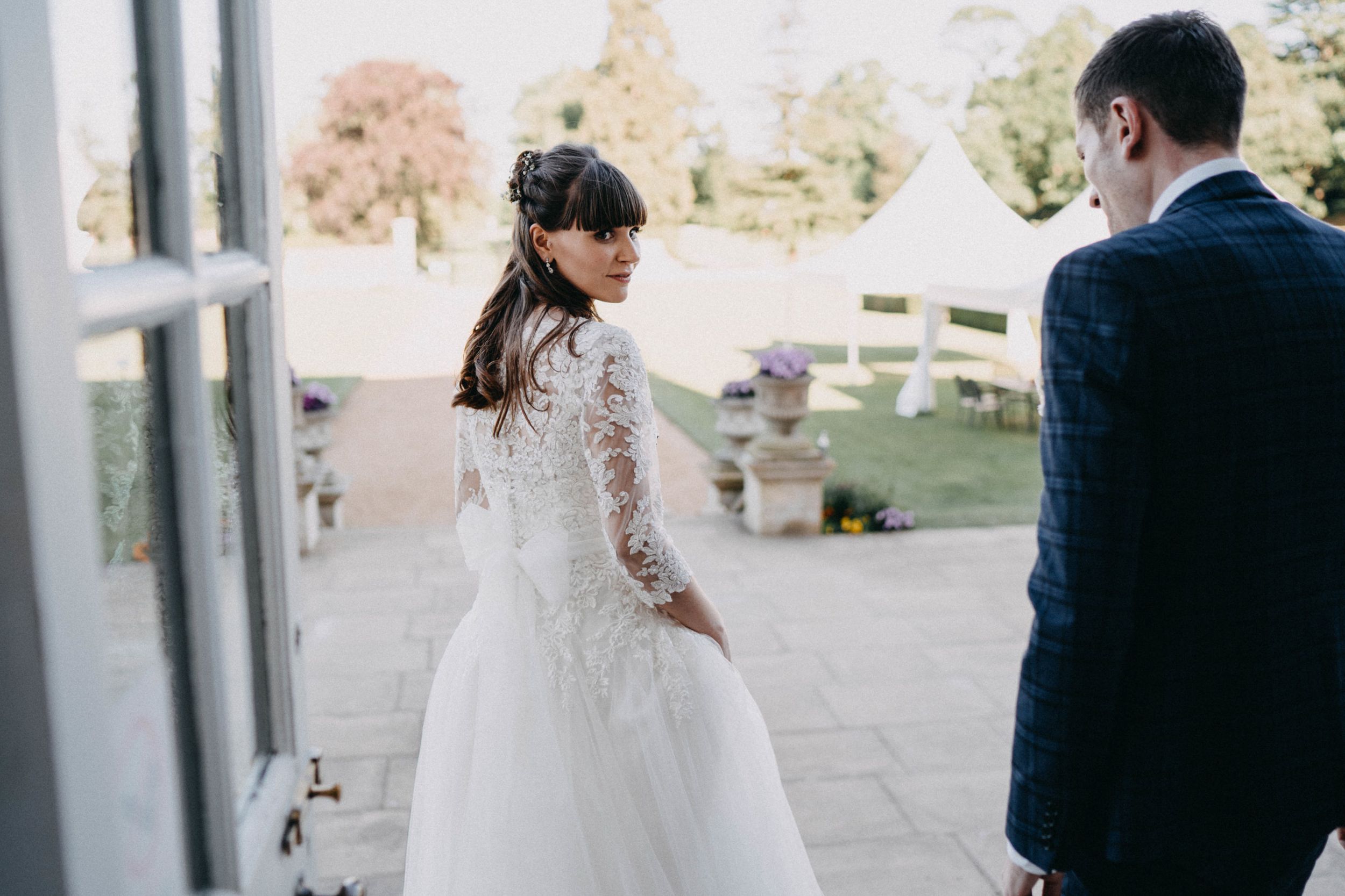 A bride in a lace-sleeved wedding gown looking over her shoulder on a sunny patio.