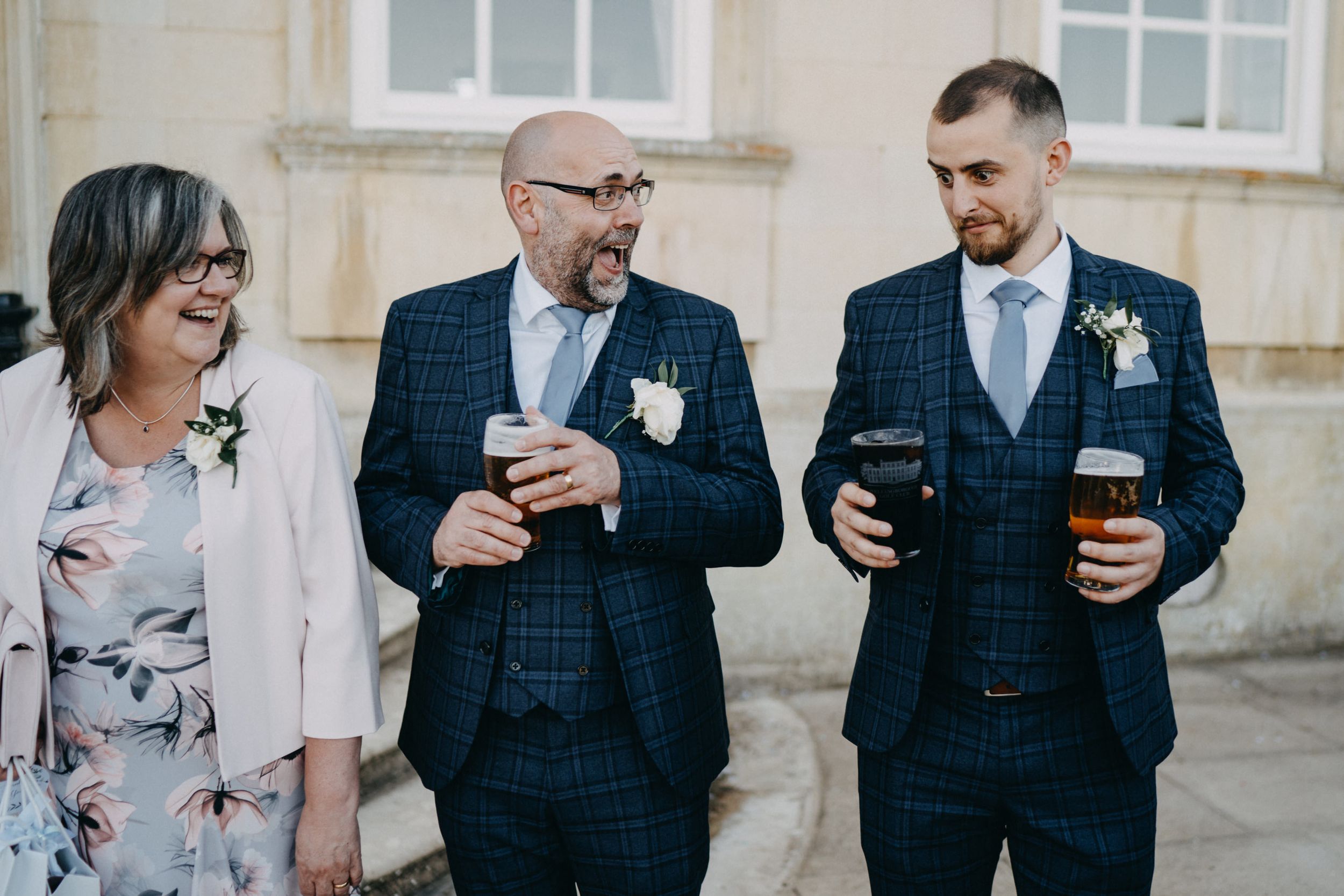 Wedding guests in formal attire share drinks and laughter outside an elegant stone building.