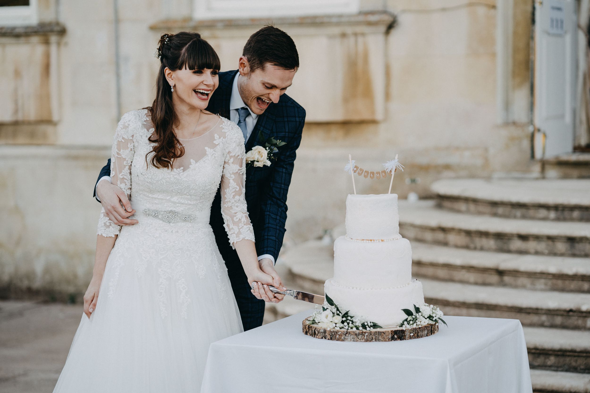 Couple cutting their elegant white wedding cake on stone steps.