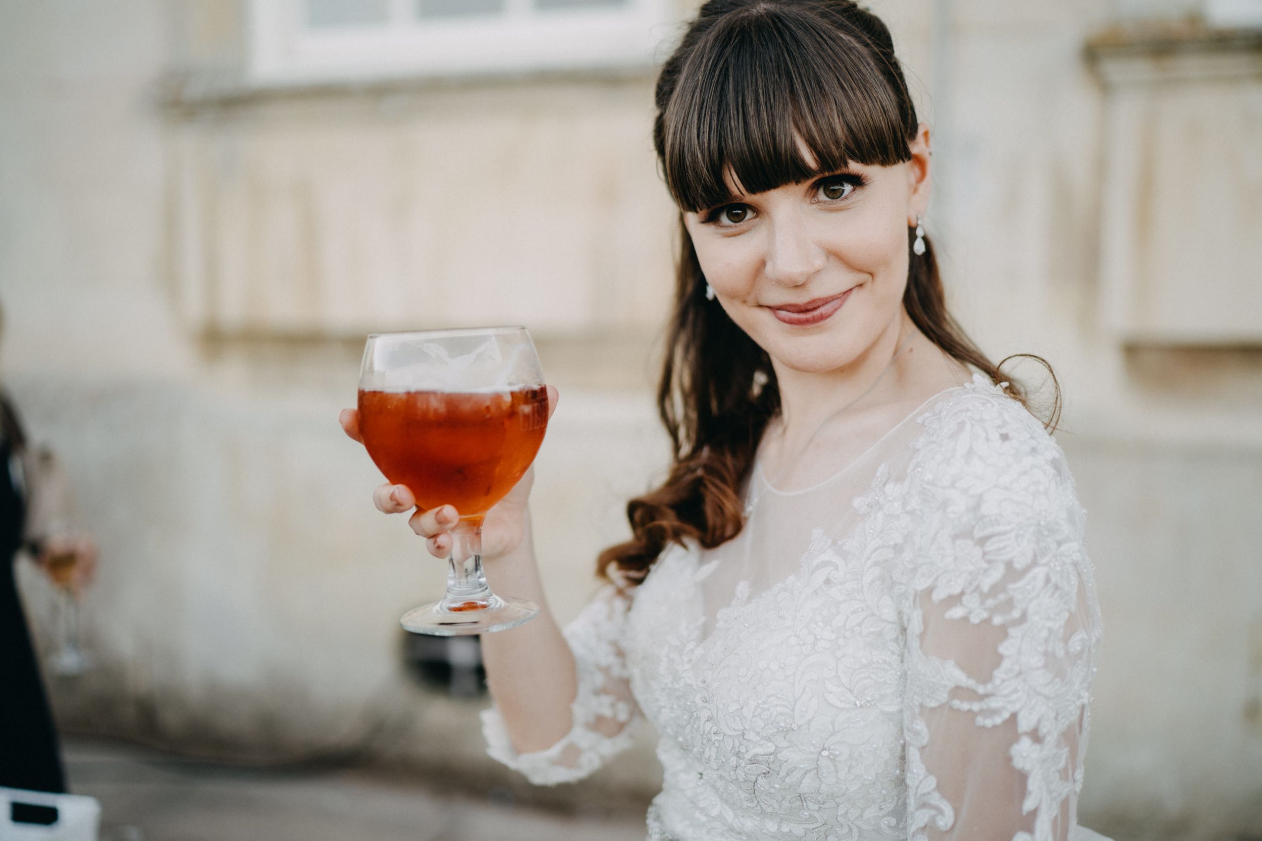 Elegant bride in lace dress holding a glass of sparkling cocktail outdoors.