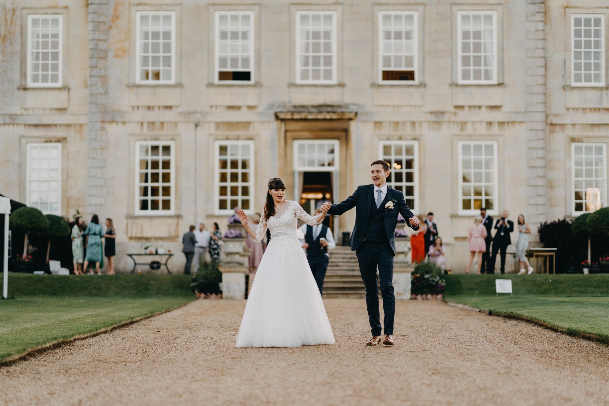 Elegant couple walking along gravel path towards historic mansion during wedding celebration at Harrowden Hall.