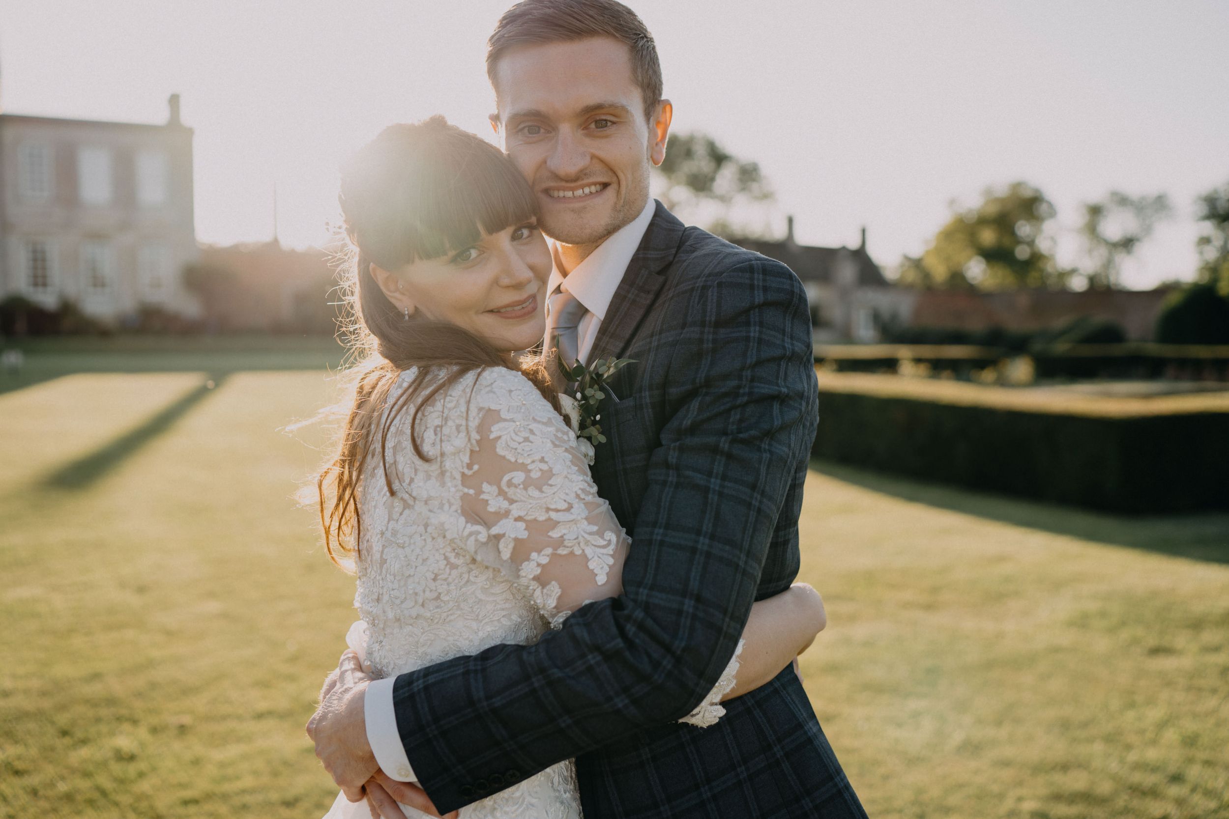 A romantic wedding couple embraces on a sunlit lawn in front of a stately mansion.