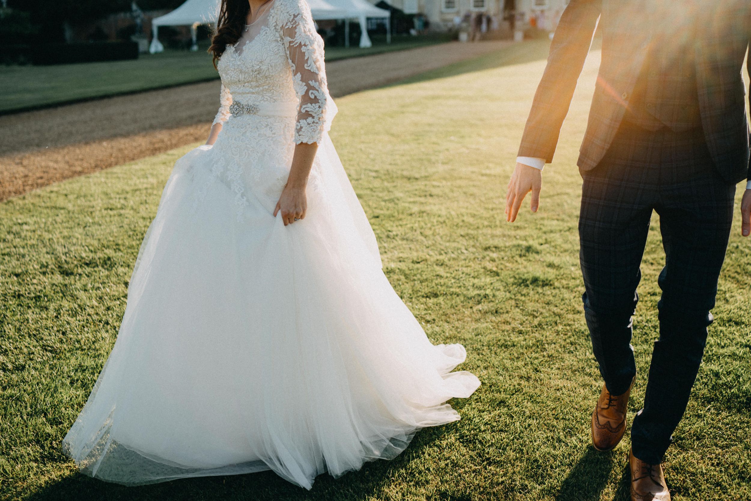 A flowing white wedding dress trails on manicured grass during an outdoor celebration.