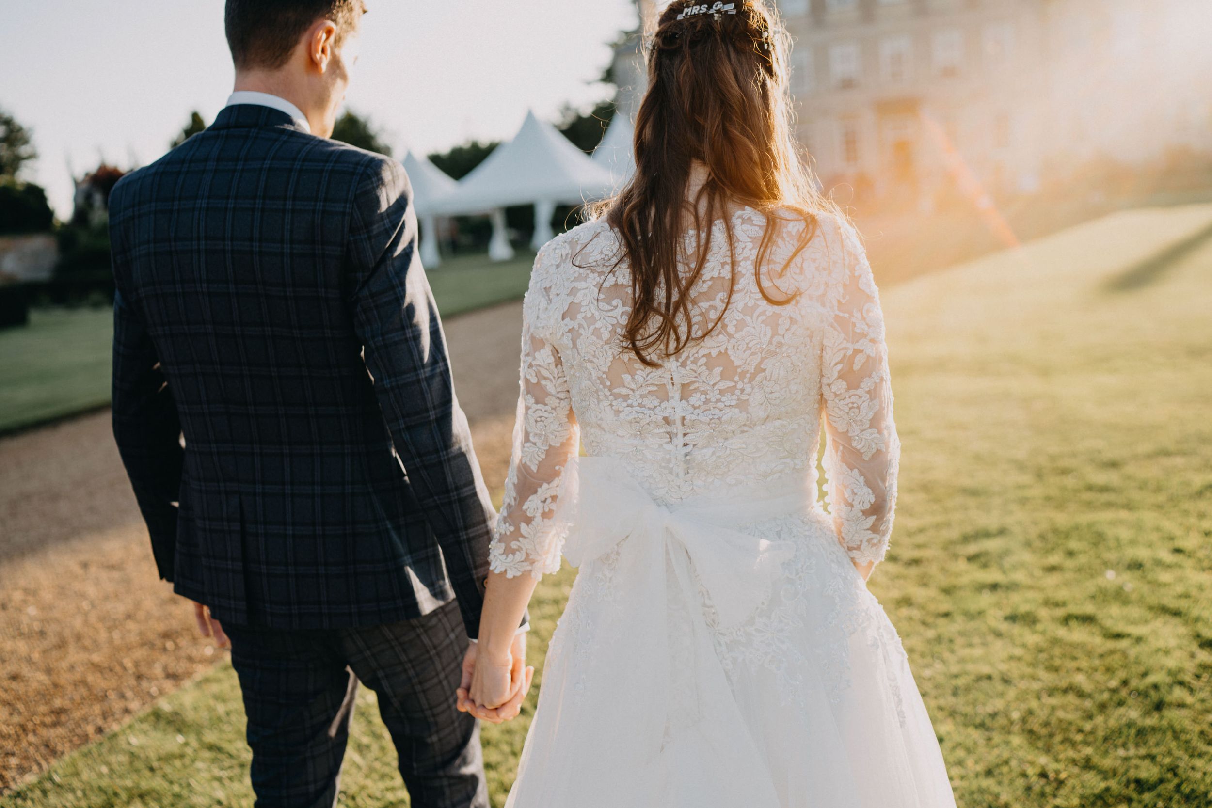 A wedding couple walks hand in hand across a sunny lawn with elegant formal attire.