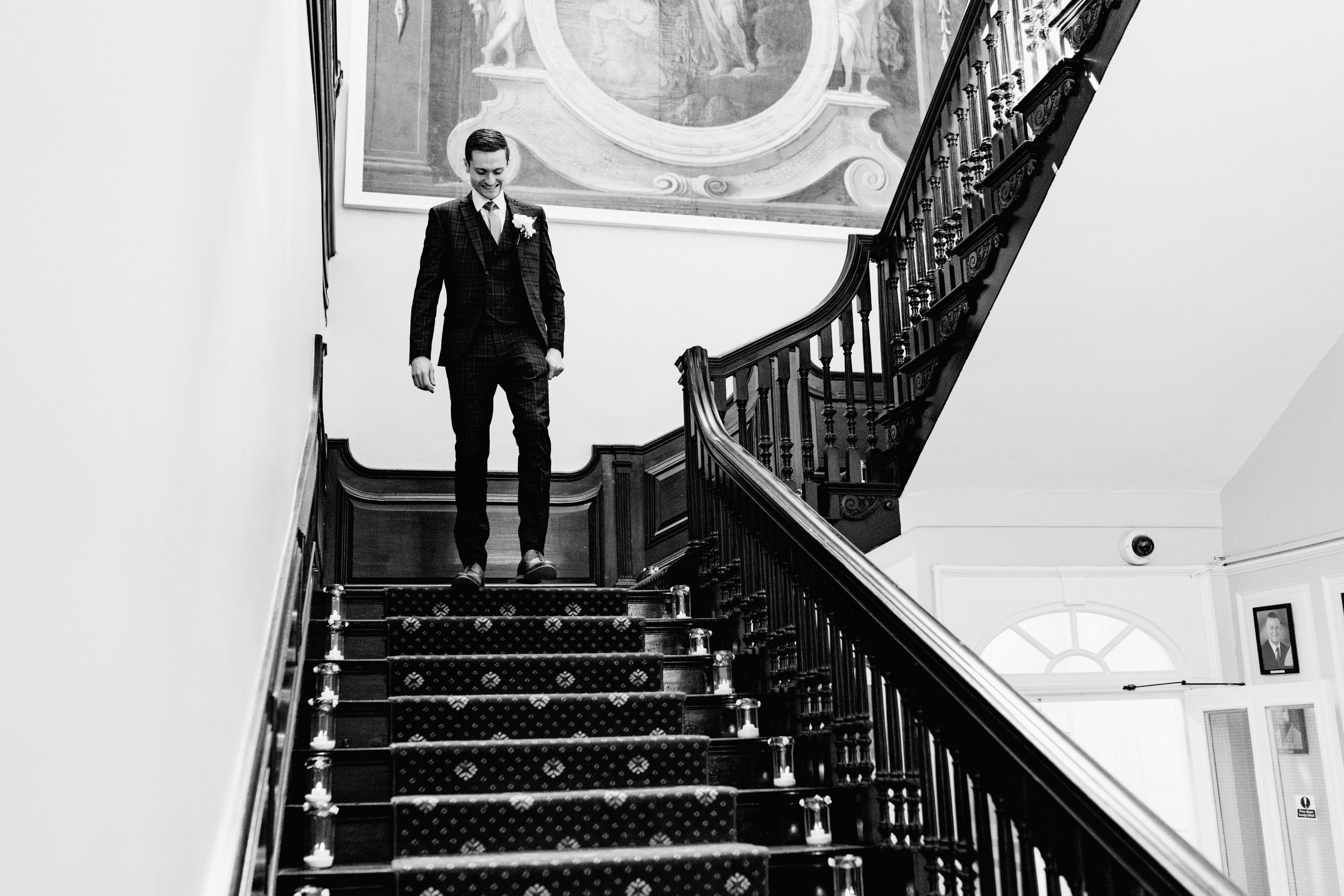 Black and white photo of the groom standing on grand staircase with ornate banister at Harrowden Hall.