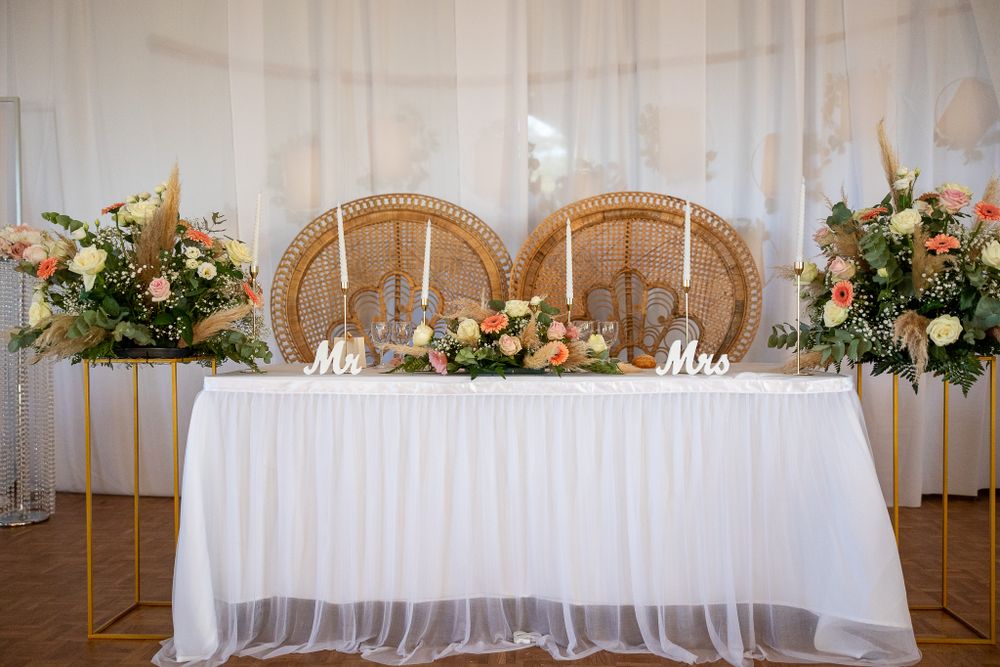 A wedding head table with wicker peacock chairs and floral arrangements against white drapery.