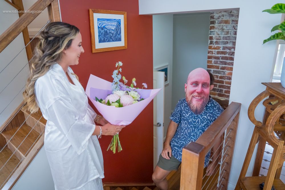 A person wearing a white robe receives a bouquet of flowers at the base of a staircase in a home interior.