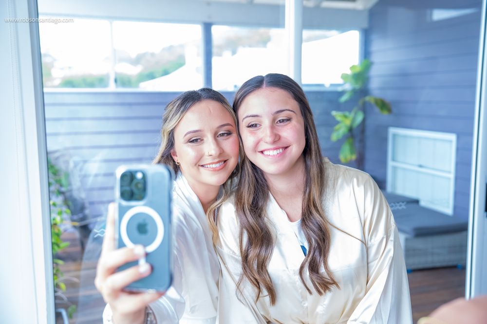 Two people in white outfits take a selfie together using a smartphone in a bright indoor setting.