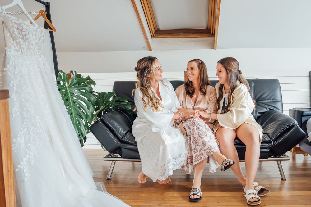 Three friends in white robes sit together on black chairs next to a hanging wedding dress.