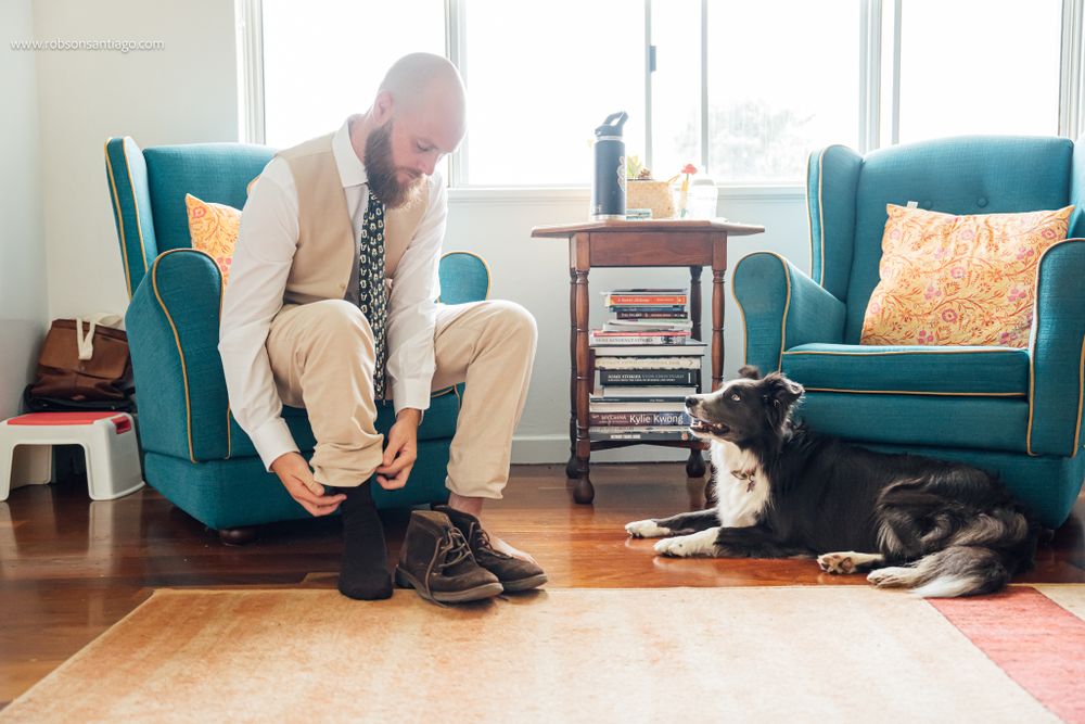 Person in casual attire putting on shoes in living room while black and white dog watches from nearby armchair.