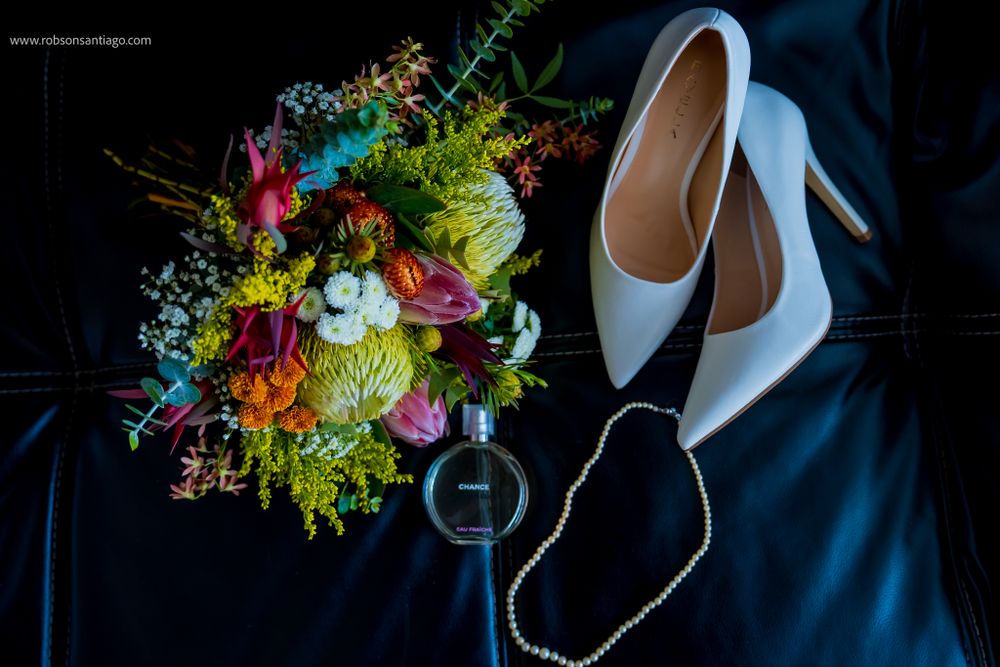 A bridal bouquet with colorful flowers next to white high heel shoes and sunglasses on dark blue fabric.