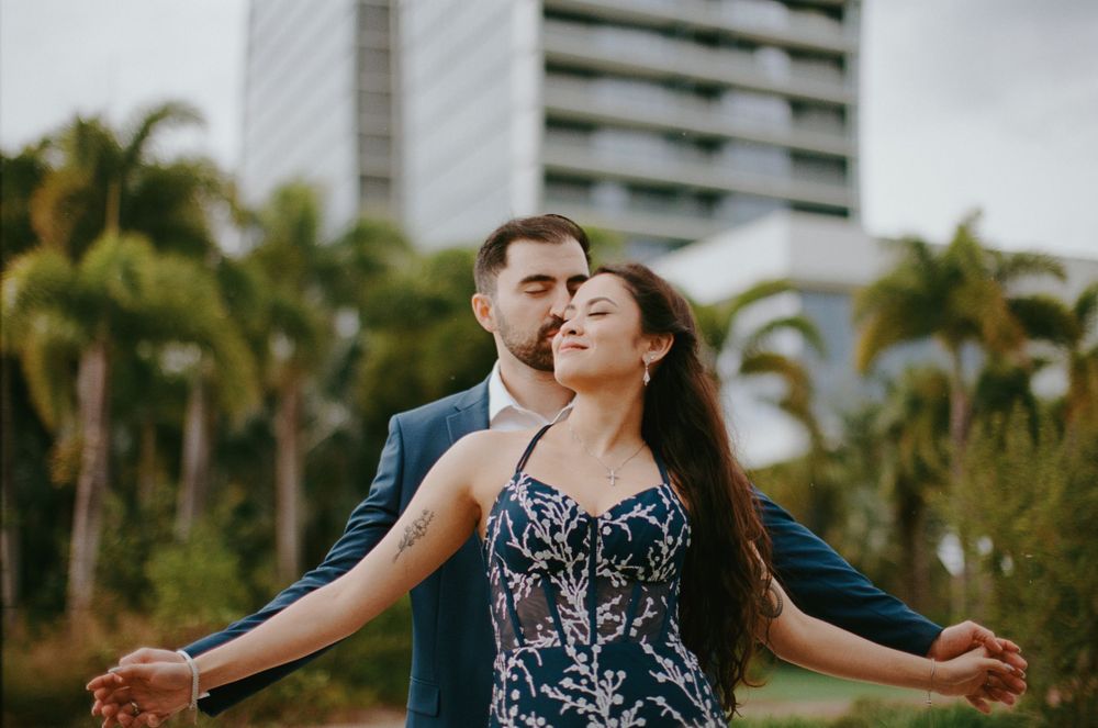 A romantic couple embraces in a lush tropical garden with palm trees and a high-rise building in the background.
