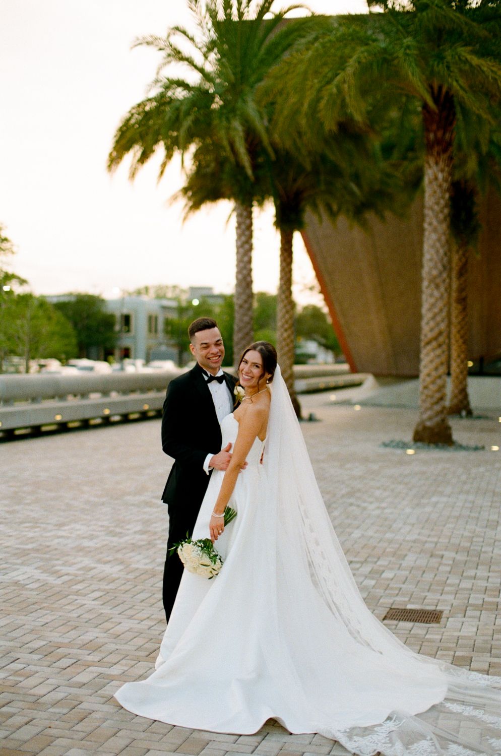 Wedding couple poses beside tall palm trees on a brick paved walkway at sunset at the Winter Park Events Center