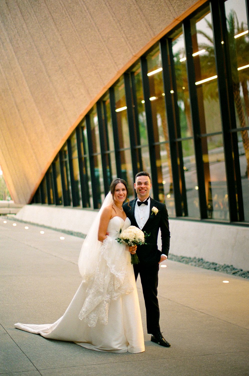 Modern wedding couple poses outside curved building with glass windows and concrete architecture in elegant attire at the Winter Park Events