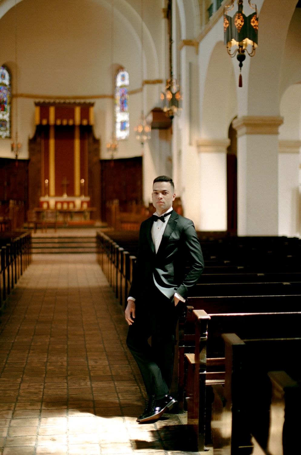 Inside a historic church sanctuary with wooden pews and arched ceiling, a person in a black tuxedo stands in formal attire.