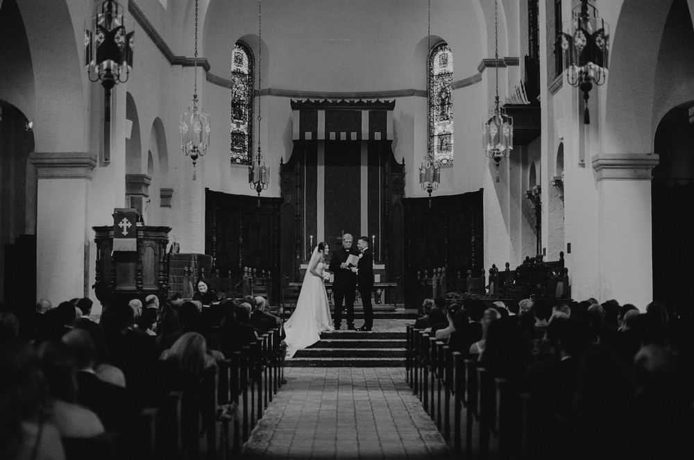 Black and white photograph at Knowles Memorial Chapel wedding ceremony with guests seated in wooden pews and figures at the altar