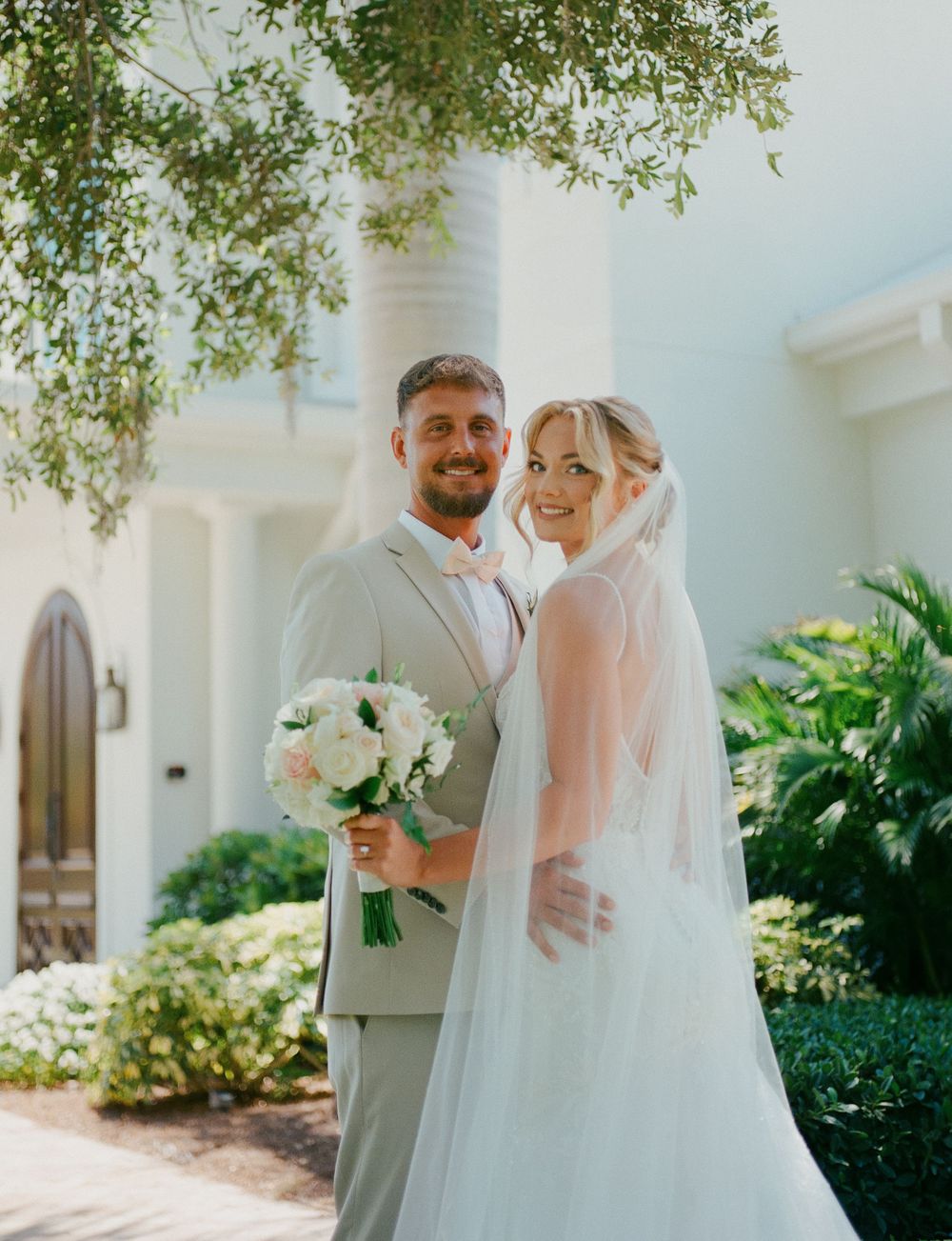 A couple poses together in traditional wedding attire with white flowers against a Mediterranean-style white building.