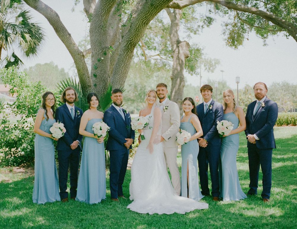 Wedding party in pale blue bridesmaid dresses and navy suits posed outdoors under large oak tree.